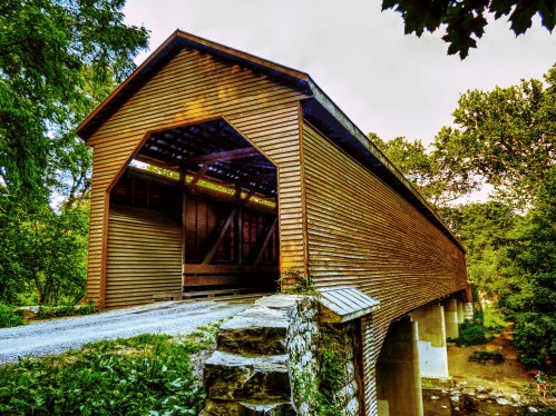 A wooden covered bridge spans a creek, surrounded by lush greenery and trees, with a gravel path leading to it.