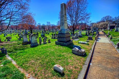 A sunny cemetery scene with gravestones and a pathway, surrounded by trees and green grass.
