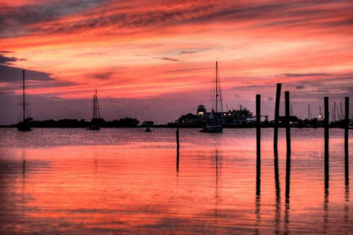 A serene sunset over a calm bay, with silhouetted boats and poles reflecting vibrant pink and orange hues in the water.