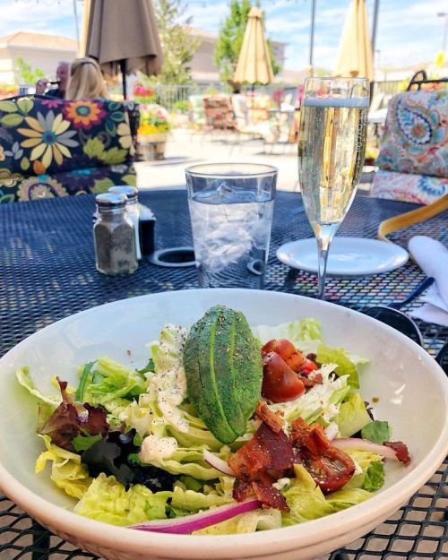 A fresh salad with bacon and avocado, served on a patio table with a glass of sparkling water and champagne.