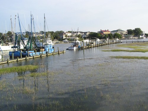 A calm waterway with fishing boats docked along a wooden pier, surrounded by marsh grass and buildings in the background.