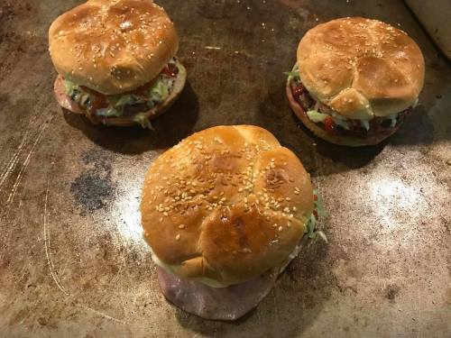 Three sesame seed buns filled with various toppings, resting on a griddle.