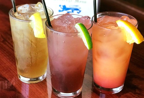 Three colorful cocktails with ice, garnished with lemon and lime, served in tall glasses on a wooden table.
