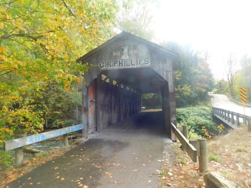 A wooden covered bridge with "C.R. Phillips" sign, surrounded by autumn foliage and a winding road nearby.
