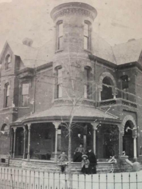 A vintage black-and-white photo of a large, ornate brick house with a turret and a porch, featuring people in front.