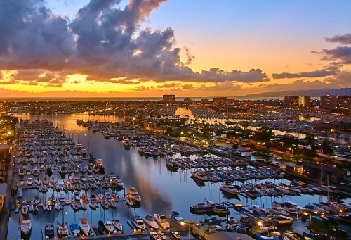 A vibrant sunset over a marina filled with boats, reflecting colorful clouds and city lights on the water.