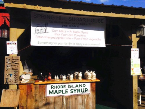A rustic stand at Spring Hill Sugar House offers maple syrup, apple cider, and fresh vegetables, with a corn maze sign.