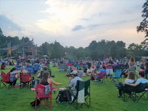 A large crowd sits on lawn chairs in a park during an outdoor event at sunset, with trees in the background.