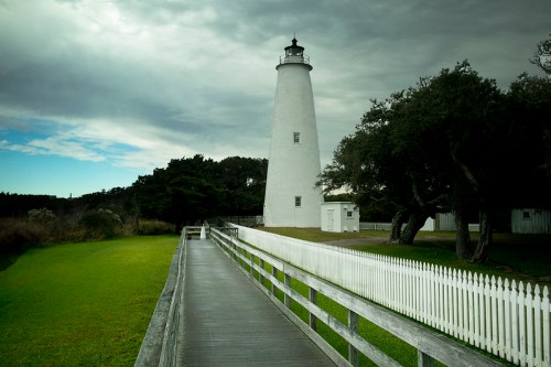 A white lighthouse stands beside a wooden walkway, surrounded by green grass and trees under a cloudy sky.