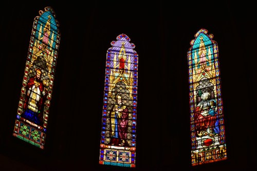 Three colorful stained glass windows depicting religious figures and scenes, illuminated against a dark background.