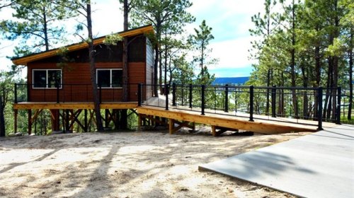 A modern cabin on stilts surrounded by trees, featuring a ramp and large windows, set in a natural landscape.