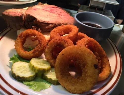A plate featuring roast beef, onion rings, pickles, and a small bowl of sauce, garnished with lettuce.