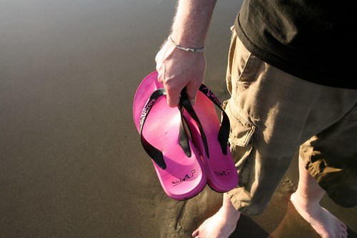 A person holding pink flip-flops while standing barefoot on a sandy beach.