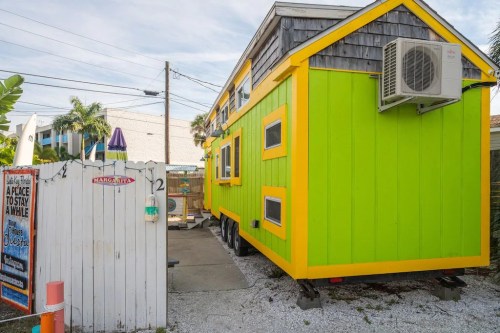 A vibrant green and yellow tiny house on wheels, surrounded by palm trees and a white picket fence.