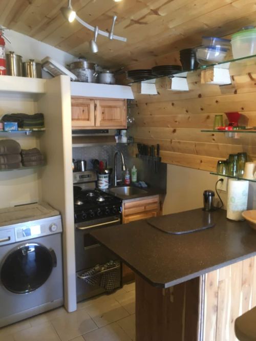 Cozy kitchen with wooden walls, equipped with a stove, sink, washing machine, and shelves of dishes and utensils.