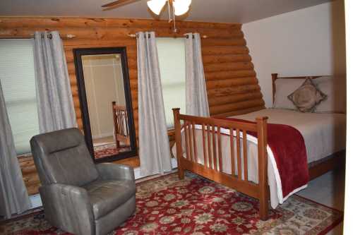 Cozy bedroom with log walls, a bed, a mirror, a chair, and a patterned rug. Natural light through window curtains.