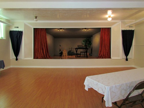 A spacious room with a stage framed by red curtains, featuring a stool, musical instruments, and a table in the foreground.