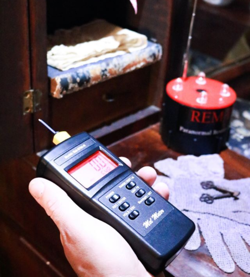 A hand holds a digital meter near a vintage cabinet, with a red device and gloves in the background.