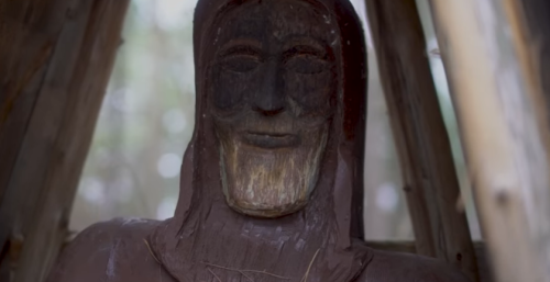 A close-up of a weathered wooden statue with a serene expression, partially sheltered by a wooden structure.