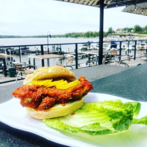 A spicy chicken sandwich with pickles on a plate, set against a waterfront view with boats and outdoor seating.