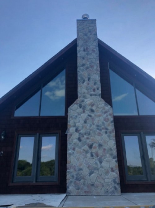 A modern A-frame house with a stone chimney and large windows, set against a clear blue sky.
