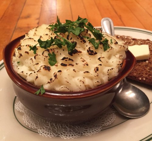 A bowl of shepherd's pie topped with browned mashed potatoes and parsley, served with a slice of buttered bread.