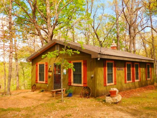 A cozy green cabin surrounded by trees, featuring a porch and red shutters, set in a tranquil outdoor setting.