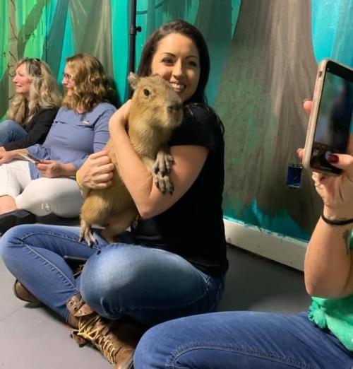 A woman smiles while holding a capybara, surrounded by friends sitting on the floor in a colorful setting.