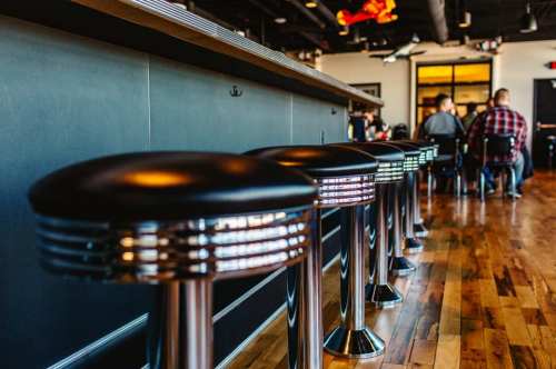 A row of black bar stools with chrome accents in a modern restaurant setting, featuring wooden floors and a bar counter.