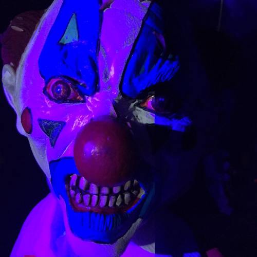A menacing clown statue with bright blue and red paint, illuminated by purple light, showcasing sharp teeth and an intense expression.