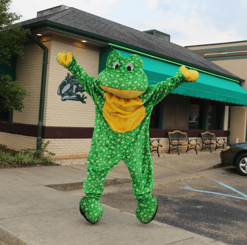A person in a green frog costume jumps joyfully outside a restaurant with a frog mural on the wall.