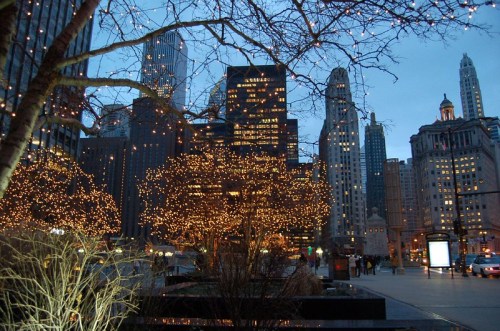 City skyline at dusk, adorned with twinkling lights on trees, creating a festive atmosphere among tall buildings.