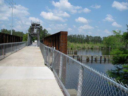 A metal bridge spans a calm river, surrounded by greenery and under a blue sky with scattered clouds.