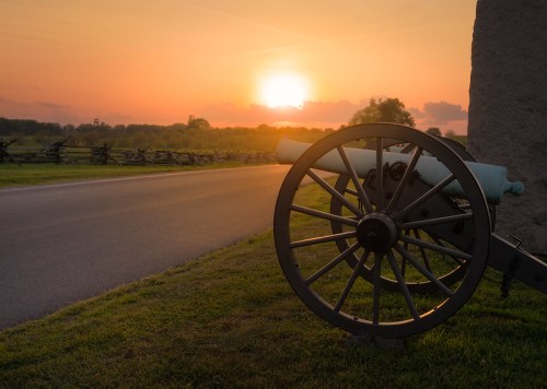 A historic cannon beside a road at sunset, with a scenic landscape and wooden fence in the background.