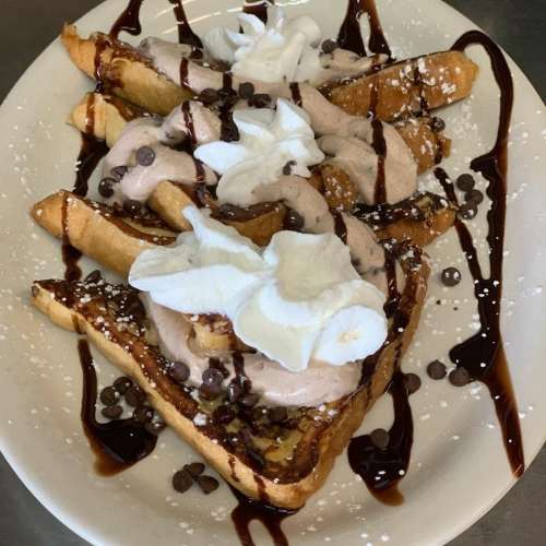 A plate of French toast topped with whipped cream, chocolate sauce, and mini chocolate chips, dusted with powdered sugar.