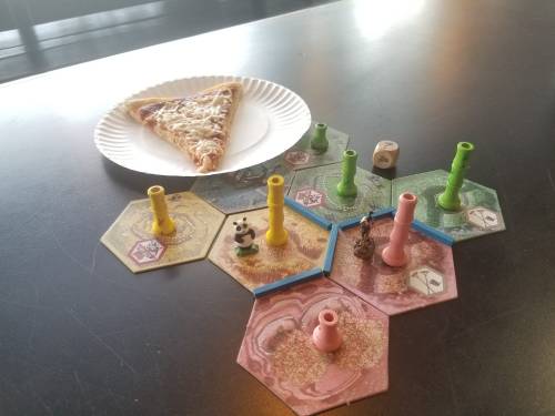 A slice of cheese pizza next to a colorful board game setup with hexagonal tiles and game pieces.