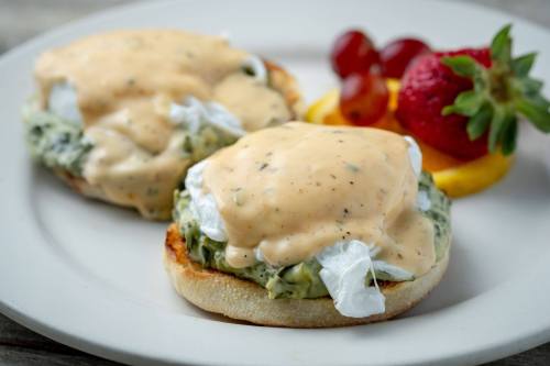 Two poached eggs on English muffins topped with creamy sauce, served with grapes and a strawberry on the side.