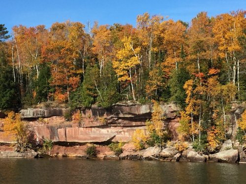 Vibrant autumn foliage on rocky cliffs beside a calm lake under a clear blue sky.