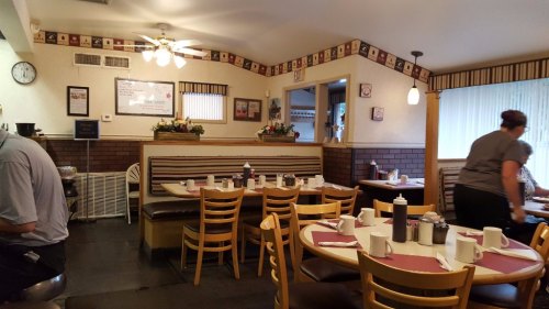 A cozy diner interior with wooden tables, chairs, and a booth, featuring floral decorations and a waitress serving.
