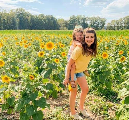 A woman carries a smiling girl on her back in a vibrant sunflower field under a sunny sky.