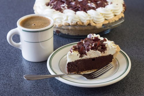 A slice of chocolate cream pie topped with whipped cream and chocolate shavings, next to a cup of coffee on a plate.