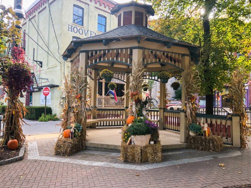 A decorated gazebo with corn stalks, pumpkins, and flowers, surrounded by autumn foliage in a quaint town setting.