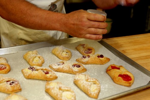 A baker sprinkles powdered sugar over freshly baked pastries on a tray.