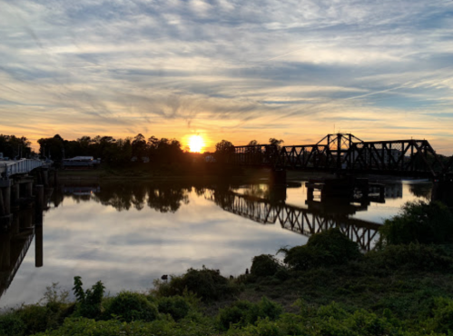 Sunset over a river with a bridge reflected in the water, surrounded by greenery and a cloudy sky.