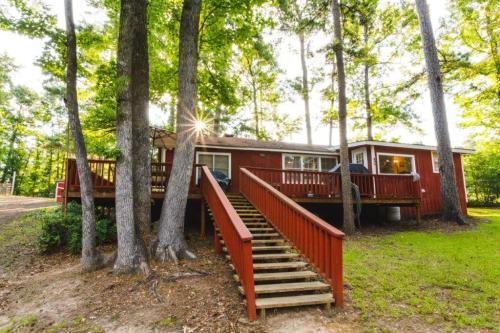 A red cabin surrounded by trees, featuring a wooden deck and stairs leading up to the entrance. Sunlight peeks through the leaves.