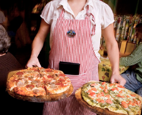 A waitress in a striped apron holds two pizzas on wooden boards in a cozy restaurant setting.
