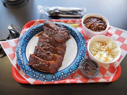 A plate of barbecue ribs with sides of coleslaw and baked beans, served on a red tray with a fork and sunglasses nearby.