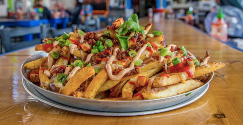 A platter of loaded fries topped with cheese, salsa, green onions, and herbs, served on a wooden table.