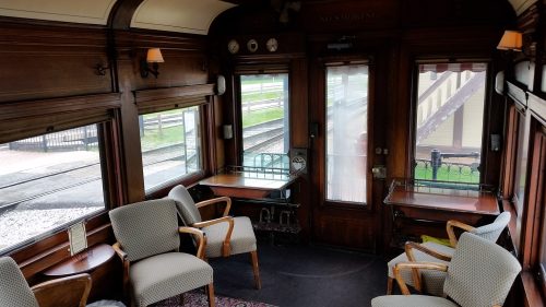Interior of a vintage train car featuring wooden paneling, plush chairs, and large windows overlooking the tracks.