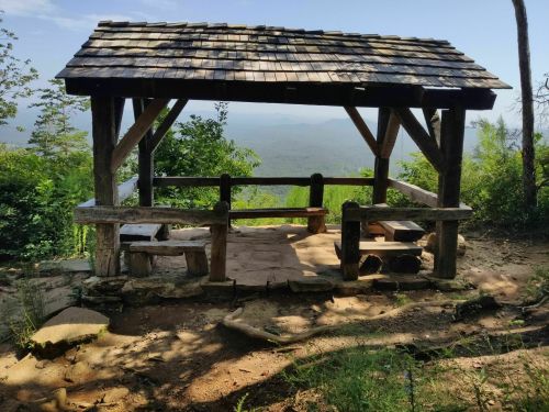 A wooden gazebo on a hillside, overlooking a scenic view of mountains and trees under a clear blue sky.